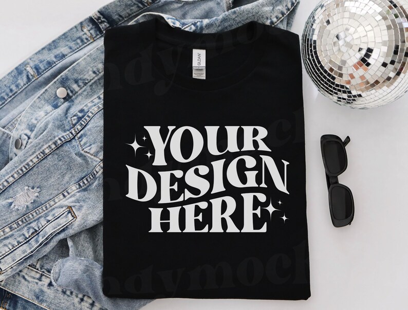 May include: A black t-shirt with white text that reads 'YOUR DESIGN HERE' with stars on either side of the text. The t-shirt is laying on a white surface with a denim jacket and a disco ball in the background.