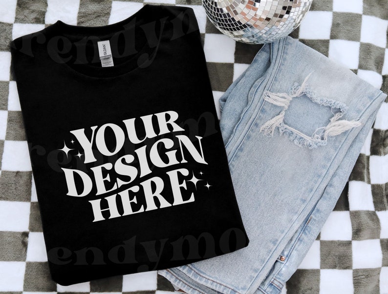 May include: A black t-shirt with the text 'YOUR DESIGN HERE' in white. The t-shirt is folded and lying on a checkered surface with a pair of blue jeans and a disco ball.