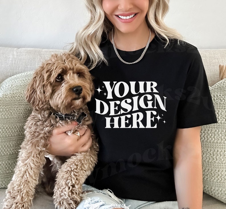 May include: A black t-shirt with the text 'YOUR DESIGN HERE' in white retro-style font. The t-shirt is being worn by a person and a brown dog is sitting on their lap.