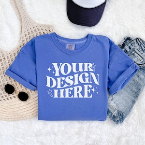 Könnte beinhalten: Ein blaues T-Shirt mit dem Text "YOUR DESIGN HERE" in Weiß, umgeben von kleinen weißen Sternen. Das Shirt liegt flach auf einer weißen, flauschigen Oberfläche.
