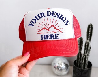 Może przedstawiać: Czerwono-biała czapka trucker z białym panelem przednim, na którym znajduje się grafika gór i napis "YOUR DESIGN HERE" w kolorze niebieskim.