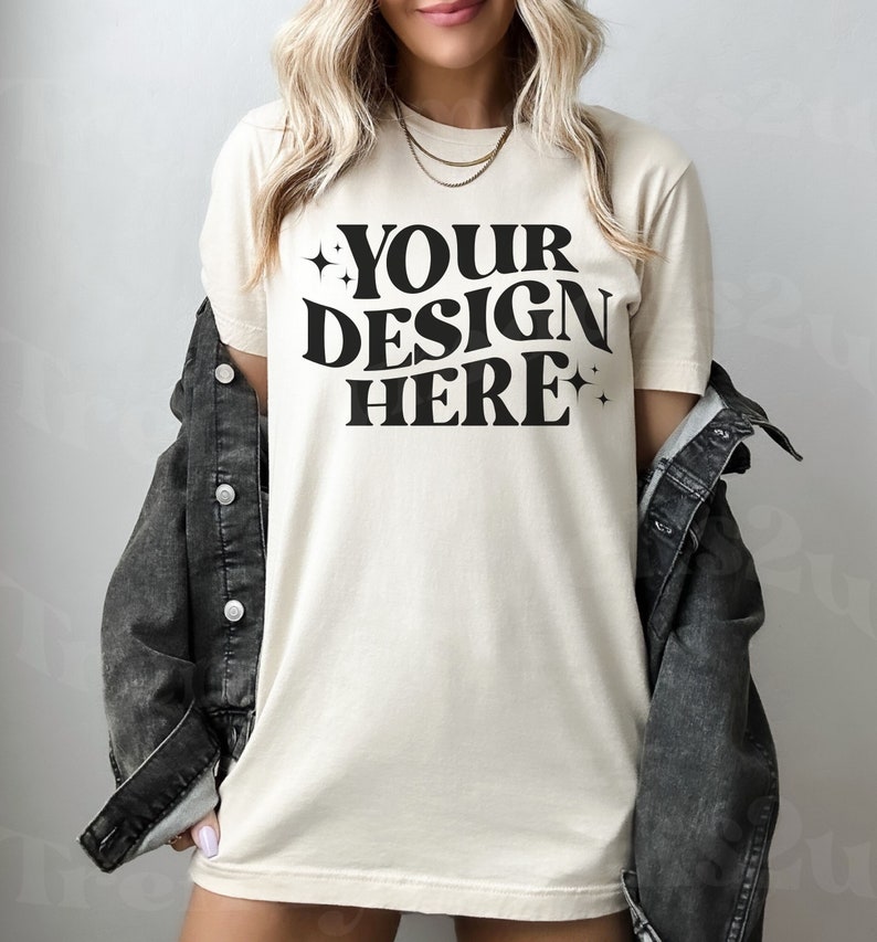 May include: A woman wearing a cream colored t-shirt with the text "YOUR DESIGN HERE" in black. She is wearing a black denim jacket and has her hair pulled back.