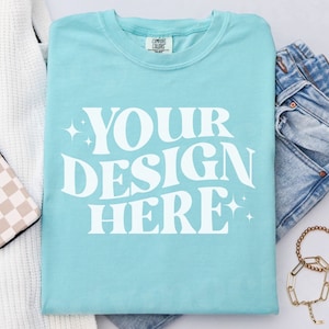 May include: A light blue t-shirt with the text "YOUR DESIGN HERE" in white. The shirt is laid flat on a white surface with a white sweater, blue jeans, and a gold bracelet.