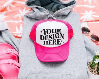 May include: A pink and white trucker hat with a black mesh back. The hat has the text "YOUR DESIGN HERE" in black letters on the front.
