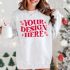 Puede incluir: Una sudadera gris con el texto "YOUR DESIGN HERE" en letras rojas. La sudadera está decorada con copos de nieve y estrellas.