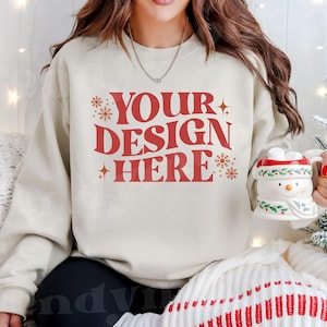 May include: A cream-colored sweatshirt with the text "YOUR DESIGN HERE" in red. The sweatshirt is decorated with snowflakes and stars. The model is wearing the sweatshirt and holding a snowman mug.