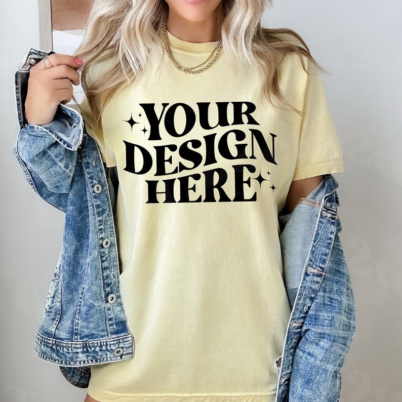May include: A pale yellow t-shirt with short sleeves and the text "YOUR DESIGN HERE" in black block letters.