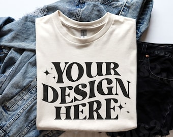 May include: A folded cream colored t-shirt with the text "YOUR DESIGN HERE" in black lettering with two small stars on either side of the text.