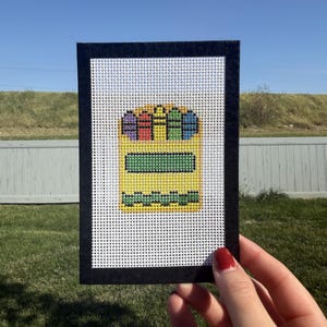 May include: A rectangular needlepoint canvas featuring a cross-stitched design of a yellow crayon box. The box contains purple, red, yellow, green, and blue crayons. It has a green rectangular label and a green wavy line at the bottom.