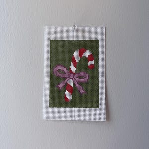 May include: A rectangular needlepoint canvas featuring a cross-stitched candy cane with red and white diagonal stripes, accented by a light purple bow. The design is centered on a solid olive green background with a white grid border.