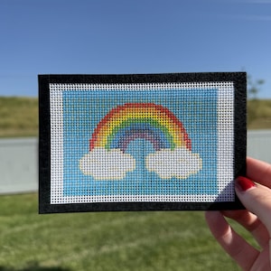 May include: A rectangular needlepoint canvas featuring a cross-stitched rainbow with red, orange, yellow, green, blue, and violet bands arched over two white clouds, all set against a light blue background and framed by a black border.