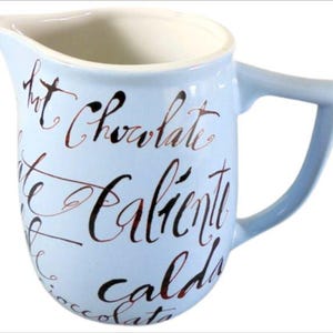 Può includere: Un creamer in ceramica blu chiaro con manico. Il creamer ha scritte nere e marroni che dicono "Hot Chocolate", "Chocolate Caliente" e "Cald".