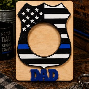 May include: A rectangular light-colored wooden picture frame featuring a central cutout shaped like a police badge. The badge design includes black and white stripes, a field of white stars, and a horizontal blue line. The word DAD is in blue.