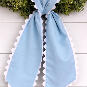 Könnte beinhalten: Hellblaues Stofftuch mit weißem, gewelltem Rand. Das Tuch ist verknotet und hängt herunter, mit einem grünen Kranz im Hintergrund. Das Tuch ist aus einem weichen Material gefertigt.