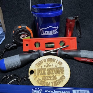 May include: A selection of DIY tools, including a blue Lowe's bucket, a tape measure, a spirit level, screwdrivers, and a wooden keychain with the text "THAT'S WHAT I DO I FIX STUFF AND KNOW THINGS". Also included is a red torch and a blue pencil.