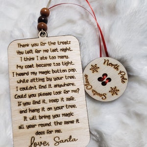 以下が含まれることがあります： サンタクロースからのメッセージが書かれた木製のクリスマスオーナメント。オーナメントはタグの形をしていて、赤いリボンが付いています。メッセージには、「昨夜、私のために残してくれたお菓子、ありがとう。たくさん食べ過ぎたみたい。コートがキツくなっちゃった。木の下に座っていたら、魔法のボタンがポンって音がしたんだ。どこにも見つからない。探してくれる？もし見つけたら、大切に保管して、木に飾ってね。それは、私と同じように、一年中魔法を届けてくれるよ。愛を込めて、サンタクロース。」と書いてあります。