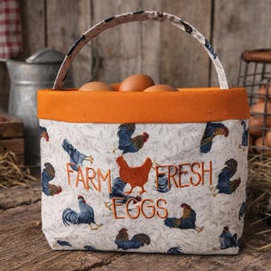 May include: A fabric egg basket with an orange rim and handle, filled with brown eggs. The basket is decorated with blue rooster illustrations and the words "FARM FRESH EGGS".