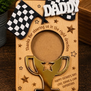 Puede incluir: Un marco de fotos rectangular de madera con un recorte de trofeo de espejo dorado, dos banderas a cuadros en blanco y negro y el texto blanco DADDY. El texto grabado dice thanks for steering us in the right direction y HAPPY FATHER'S DAY! LOVE, SOPHIE & LIAM SUPERSTARS.