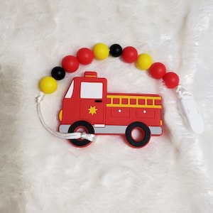 May include: A red fire truck silicone teether with a white string and a black and yellow beaded chain. The teether has a white clip attached.