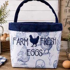 May include: A fabric basket with a dark blue handle and trim, featuring a white exterior printed with blue illustrations of chickens and nests. The text "FARM FRESH EGGS" is embroidered in dark blue thread across the centre of the basket.