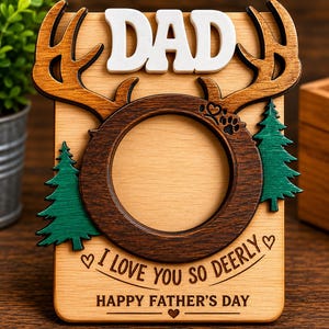May include: A rectangular wooden photo frame featuring a white DAD cutout at the top, a dark brown circular center with deer antlers, and green pine trees on the sides. The text reads I LOVE YOU SO DEERLY and HAPPY FATHER'S DAY with heart icons.