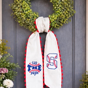 May include: A green wreath hangs on a gray door, with a white banner featuring baseball-themed embroidery. The banner has red scalloped edges and the text "tis the season" and a baseball-style number 5.