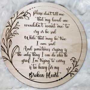 May include: A round wooden sign with a floral design and the text "Please don't tell me that my loved one wouldn't want me to cry or be sad. While that may be true I am sad. And sometimes crying is the only thing I can do when the grief I'm trying to carry is too heavy for my Broken Heart."