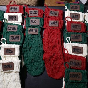 May include: A collection of 18 personalized Christmas stockings with names on brown leather tags. The stockings are made of knitted fabric in red, white, and green.