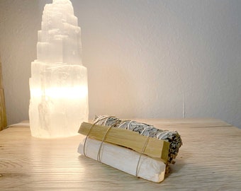 Può includere: Una lampada di selenite bianca e luminosa è posizionata accanto a un fascio di salvia e bastoncini di palo santo, legati con spago. La salvia è grigio-verde e il palo santo è marrone chiaro. Gli oggetti sono su una superficie di legno chiaro.