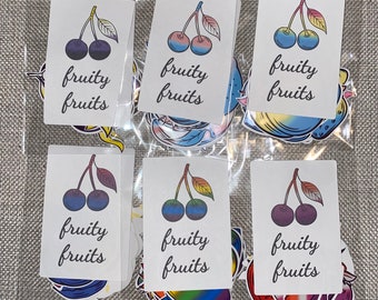 May include: A collection of colorful stickers in a clear plastic bag. Each sticker features a pair of cherries with leaves and the words "fruity fruits." The cherries are in various colors, including blue, purple, and rainbow hues.
