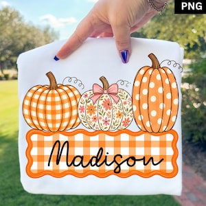 May include: A white t-shirt featuring a graphic of three pumpkins above a scalloped orange and white gingham banner with the name Madison written in black script. The left pumpkin has an orange gingham pattern, the center has a floral print with a pink bow, and the right pumpkin is orange with white polka dots.