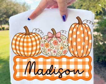 May include: A white t-shirt featuring a graphic of three pumpkins above a scalloped orange and white gingham banner with the name Madison written in black script. The left pumpkin has an orange gingham pattern, the centre has a floral print with a pink bow, and the right pumpkin is orange with white polka dots.