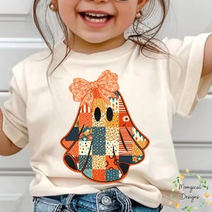 Könnte beinhalten: Cremefarbenes T-Shirt mit einem Patchwork-Geister-Design und einer großen orangefarbenen Schleife. Der Geist besteht aus verschiedenen gemusterten Quadraten in Orange, Blau und Gelb. Geeignet für Halloween oder Herbst.