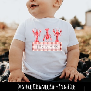 May include: White baby t-shirt with three red lobsters and a red and white checkered banner with the name "JACKSON" printed on it.
