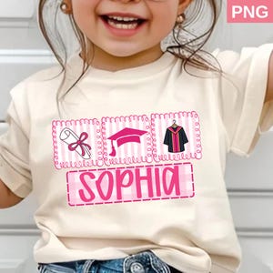 May include: A cream-colored t-shirt featuring a graduation-themed design. The design includes a diploma, graduation cap, and gown, all in pink, with the name "SOPHIA" in a pink, block letter font. The shirt is worn by a young child.