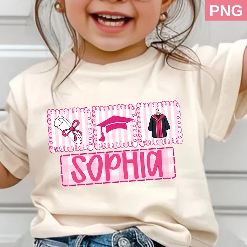 May include: A cream-colored t-shirt featuring a graduation-themed design. The design includes a diploma, graduation cap, and gown, all in pink, with the name "SOPHIA" in a pink, block letter font. The shirt is worn by a young child.