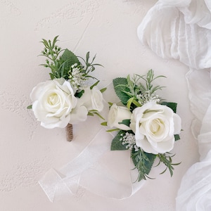 May include: Two white rose boutonnieres with greenery and white ribbon. The flowers are accented with small white buds and green leaves. The stems are wrapped in brown twine. The background is a light, textured surface with white fabric.