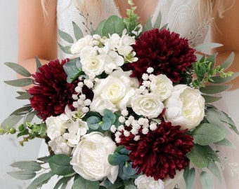 Op de afbeelding: Een bruid in een witte trouwjurk houdt een boeket bloemen vast. Het boeket bevat bordeauxrode dahlia's, witte rozen en groen. De bruid heeft lang blond haar en staat voor een witte muur met textuur.