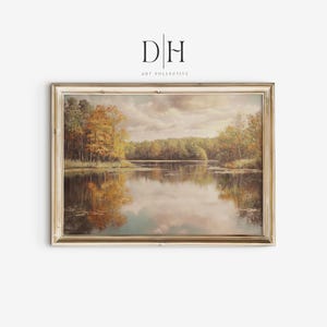 May include: A framed landscape painting depicts a serene lake scene with trees in autumn colors. The artwork features a reflective lake mirroring the sky and foliage, framed in a gold-toned border. The art is from DH Art Collective.