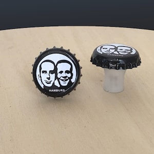 May include: Two black bottle caps with white images of two men's faces and the word "HAMBURG" printed on them. One bottle cap is on a wooden surface and the other is on a white plastic cap.