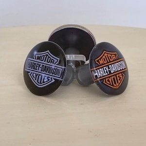 May include: Three black round motorcycle handlebar end caps with the Harley-Davidson logo in silver and orange.