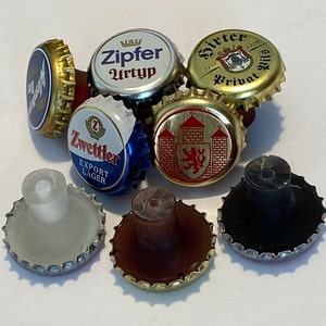 May include: Assortment of seven bottle caps, some with logos and text, including "Zipper Urtyp", "Zwettler Export Lager", and "Hirter Privat 1270", and some with a plain black, brown, or white surface. The bottle caps are arranged on a white surface.