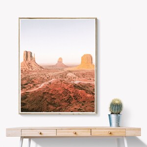 Puede incluir: Impresi&oacute;n enmarcada del paisaje de Monument Valley, con sus emblem&aacute;ticos buttes. La foto presenta tonos c&aacute;lidos de rojo y naranja, bajo un cielo azul p&aacute;lido. La impresi&oacute;n se exhibe en un escritorio de madera clara con un peque&ntilde;o cactus.