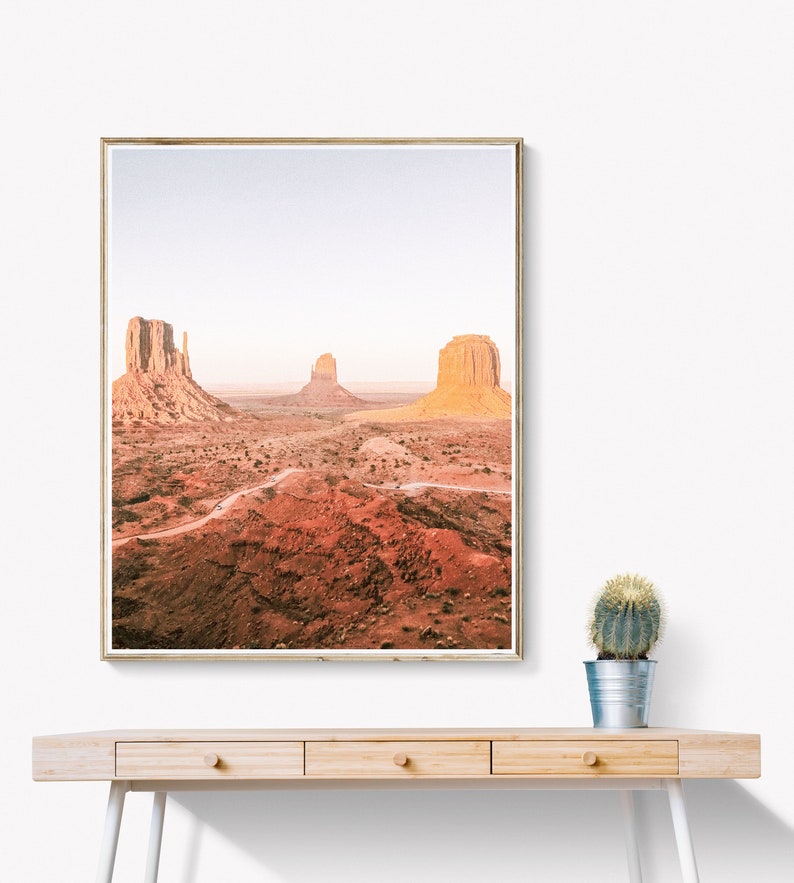 Puede incluir: Impresi&oacute;n enmarcada del paisaje de Monument Valley, con sus emblem&aacute;ticos buttes. La foto presenta tonos c&aacute;lidos de rojo y naranja, bajo un cielo azul p&aacute;lido. La impresi&oacute;n se exhibe en un escritorio de madera clara con un peque&ntilde;o cactus.
