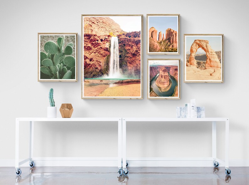 Puede incluir: Una colecci&oacute;n de impresiones enmarcadas de paisajes des&eacute;rticos, que incluyen una cascada, un cactus y un arco natural. Las impresiones se exhiben en una mesa rodante blanca, creando una est&eacute;tica de pared de galer&iacute;a.