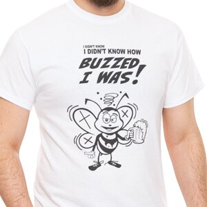 May include: White t-shirt with a black and white cartoon bee design. The bee is holding a beer mug and has a dizzy expression. The text on the shirt reads "I didn't know how buzzed I was!"