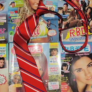 May include: A red and white striped tie with blue stripes. The tie is lying on top of several magazines with the word "Rebelde" on the covers. The magazines are in Spanish.