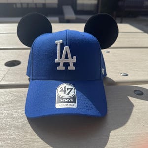 May include: A blue baseball cap with black Mickey Mouse ears. The cap has a white "LA" logo on the front and a white "47 MVP ADJUSTABLE" label on the side.