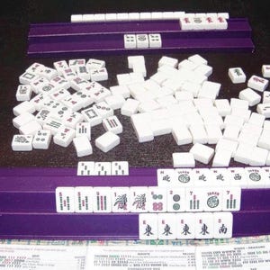 K&ouml;nnte beinhalten: Ein Mahjong-Spiel mit wei&szlig;en rechteckigen Spielsteinen, die schwarze, rote und gr&uuml;ne Symbole, Schriftzeichen und Zahlen aufweisen. Die Steine sind auf zwei violetten Kunststoffhaltern angeordnet, mit einem Haufen Steine in der Mitte.