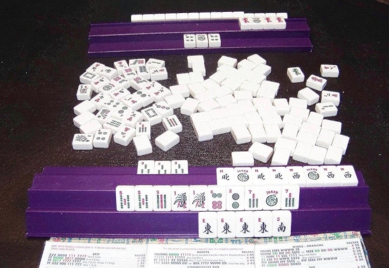 K&ouml;nnte beinhalten: Ein Mahjong-Spiel mit wei&szlig;en rechteckigen Spielsteinen, die schwarze, rote und gr&uuml;ne Symbole, Schriftzeichen und Zahlen aufweisen. Die Steine sind auf zwei violetten Kunststoffhaltern angeordnet, mit einem Haufen Steine in der Mitte.